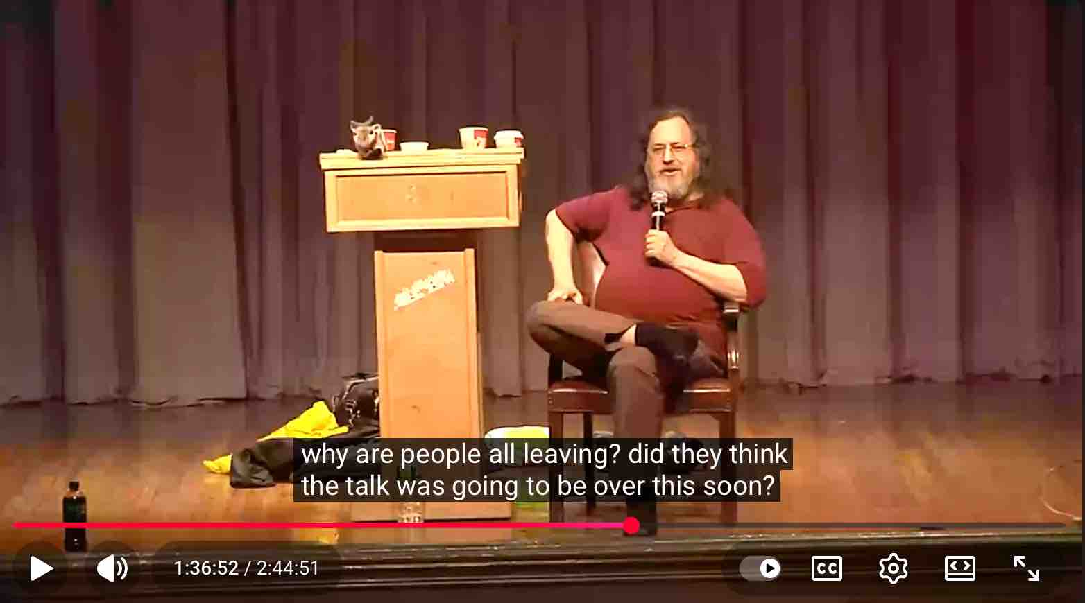 a screenshot from a youtube video of richard stallman speaking in front of an audience; he is sitting in a chair, and has no shoes on. the subtitle reads "why are people all leaving? did they think the talk was going to be over this soon?" the video timestamp shows we are 1 hour 36 minutes into a 2 hour, 44 minute speech.